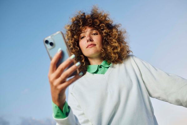 Portrait of a curly brunette checking her smartphone while standing in front of a clear sky.